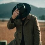 A man in a fashionable coat and hat poses stylishly outdoors against a scenic backdrop.