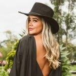 Chic woman with blonde hair wearing a black fedora outdoors, showcasing a backless outfit and confident pose.